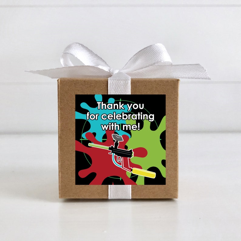 May include: A small brown box with a white ribbon tied around it. The box has a square sticker on the front with a black background, colourful paint splatters, a paintball gun, and the text "Thank you for celebrating with me!"