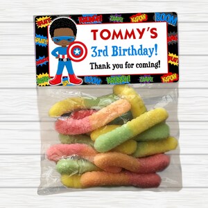 May include: A party favor bag with a superhero theme. The bag has a label with a cartoon superhero and the text "TOMMY'S 3rd Birthday! Thank you for coming!" The bag contains colorful gummy worms.