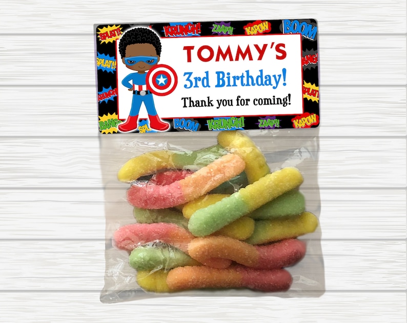 May include: A party favor bag with a superhero theme. The bag has a label with a cartoon superhero and the text "TOMMY'S 3rd Birthday! Thank you for coming!" The bag contains colorful gummy worms.