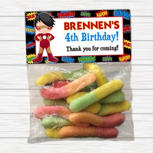 May include: A superhero-themed birthday party favor bag with a red, yellow, and blue cartoon superhero on the front. The bag has the text "BRENNEN'S 4th Birthday! Thank you for coming!" printed on it. The bag is filled with colorful gummy worms.