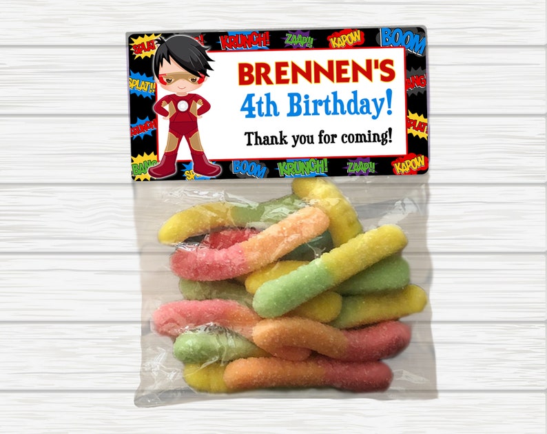 May include: A superhero-themed birthday party favor bag with a red, yellow, and blue cartoon superhero on the front. The bag has the text "BRENNEN'S 4th Birthday! Thank you for coming!" printed on it. The bag is filled with colorful gummy worms.