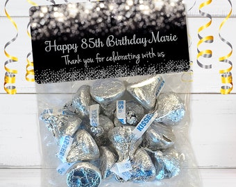 Puede incluir: Una bolsa de regalo negra y plateada con una etiqueta que dice "Feliz cumpleaños número 85 Marie Gracias por celebrar con nosotros". La bolsa está llena de besos de chocolate envueltos en papel de aluminio plateado.