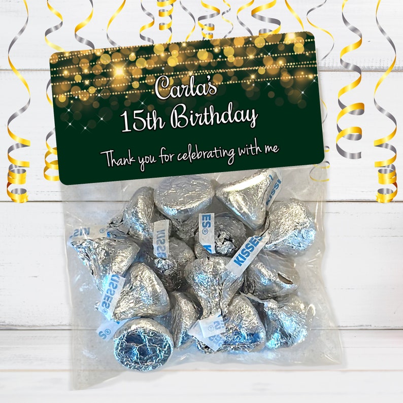 Puede incluir: Una bolsa de regalo verde y dorada con una etiqueta personalizada que dice "Carla's 15th Birthday. Thank you for celebrating with me." La bolsa est&aacute; llena de besos de chocolate envueltos en papel plateado.
