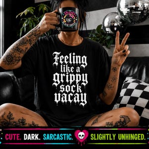 May include: A black t-shirt featuring white gothic-style text that reads Feeling like a grippy sock vacay. The shirt is paired with a black mug displaying a graphic of a black sheep with the text The Snarky Black Sheep printed on the front.