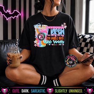 May include: A black t-shirt featuring a colorful graphic of a coffee cup with the text "she's drinkin 7 brew i can tell by the way she walks into the room" and a "Seven Brew" logo. The design includes pink, blue, and orange swirls with star accents.