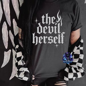 May include: Black t-shirt with the text "the devil herself" in white gothic lettering. The shirt is paired with a black and white checkered cardigan. The image also features a logo for "The 11th Black Sheep".