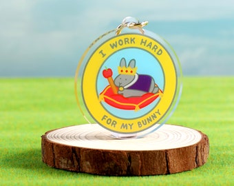 May include: A round, clear acrylic keychain with a yellow border and a blue center. The center features a cartoon gray rabbit wearing a purple robe and a gold crown, holding an apple on a stick. The text around the image reads "I work hard for my bunny".