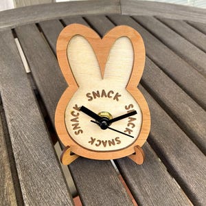 May include: Wooden bunny-shaped clock with the word "SNACK" in a circle around the clock face. The clock has black hands and a gold-colored center. The clock is made of light brown wood and sits on a wooden surface.
