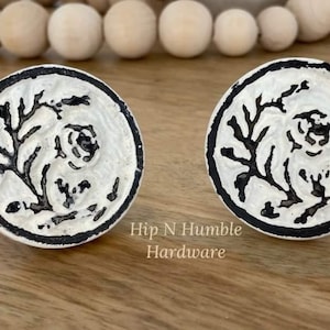 May include: Two white and black cabinet knobs with a floral design. The knobs are circular with a raised floral pattern and a black border. The text "Hip N Humble Hardware" is visible. Ideal for furniture.