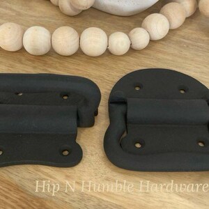May include: Two black metal drawer pulls in a vintage style. One pull is a simple rectangular shape with a handle, while the other is rounded with a loop handle.
