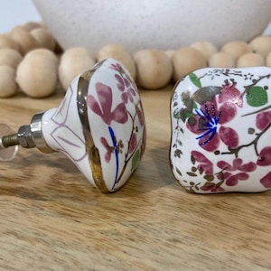 May include: Two square ceramic cabinet knobs with a white background and pink floral design. The knobs have a gold trim and a screw on the back.