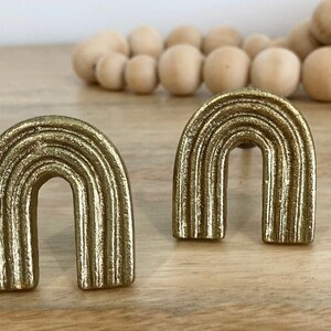 May include: Two gold-coloured metal rainbow-shaped drawer pulls. The pulls have a textured, ribbed surface.
