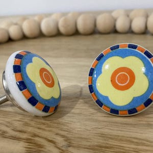 May include: Two ceramic drawer knobs featuring a blue, orange and yellow flower design. The knobs have a silver screw for attaching to a drawer.
