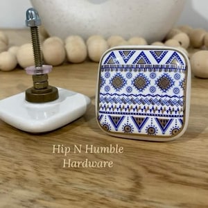 May include: Square ceramic cabinet knob with a white background and a blue and gold geometric pattern. The knob has a screw and a metal base.