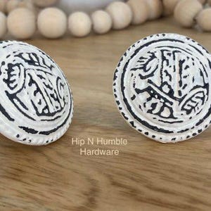 May include: Two white ceramic cabinet knobs with a black, intricate design. The knobs are round and have a raised edge. The text "Hip N Humble Hardware" is visible below the knobs.
