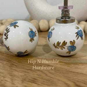 May include: Two white ceramic cabinet knobs with a blue and gold floral design. The knobs are round and have a silver metal base.