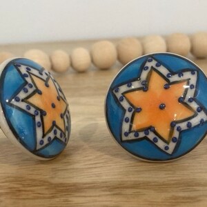 May include: Two ceramic drawer knobs with a blue background and a yellow star design. The star has a white outline and blue dots. The knobs are ideal for adding a touch of whimsy to any room.