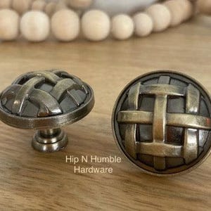May include: Two antique brass cabinet knobs with a woven design. The knobs are round and have a raised, textured pattern. The knobs are on a wooden surface.