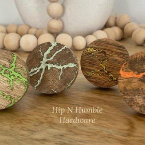 May include: Four round wooden knobs with a natural wood finish and green, gold, and orange paint accents. The knobs are arranged in a semi-circle on a wooden surface. Hip N Humble Hardware is printed below the knobs.