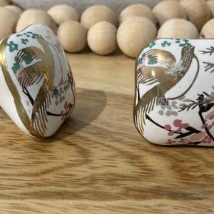 May include: Two white ceramic cabinet knobs with gold accents. The knobs feature a floral design with birds and leaves.