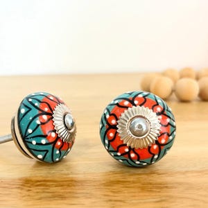 May include: Two round ceramic cabinet knobs featuring a teal and orange floral pattern with white dots and black line details. Each knob has a silver-coloured metal rosette at the centre and a threaded metal bolt extending from the back side.