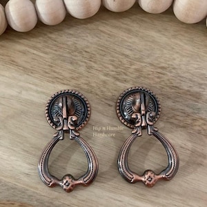 May include: Two antique copper drawer pulls with a decorative ring design. The pulls have a dark copper finish and a detailed ornate design.