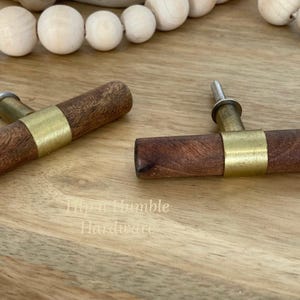 May include: Two wooden cabinet knobs with a brass accent. The knobs have a dark brown wood finish and a brass band around the centre. The knobs are shown on a light wood surface.