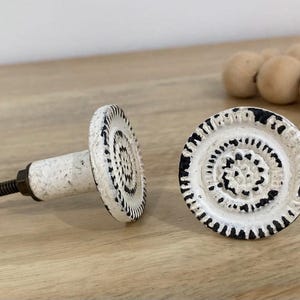 May include: Two white and black decorative cabinet knobs with a circular design. The knobs have a distressed finish and a raised circular pattern.