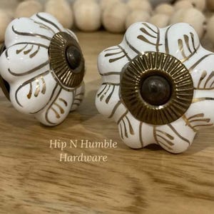 May include: Two white ceramic cabinet knobs with gold accents and a brass base. The knobs are shaped like a flower with a raised centre. The text "Hip N Humble Hardware" is visible below the knobs.