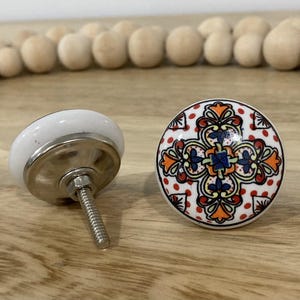 May include: Two ceramic cabinet knobs with a white base and a silver metal screw. One knob has a white base with a silver metal screw. The other knob has a colourful floral design with red, orange, yellow, blue, and white colours.
