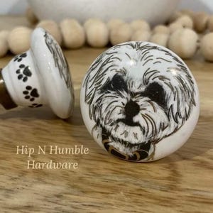 May include: Two white ceramic door knobs with black paw prints and a black and white dog illustration. The dog is wearing a gold collar. The text "Hip N Humble Hardware" is printed on the wooden surface.