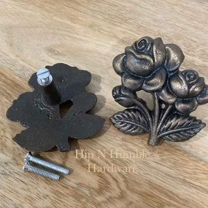 May include: Two antique bronze-coloured rose-shaped drawer pulls with a screw for installation. The pulls are on a wooden surface. Hip N Humble Hardware.