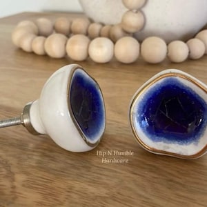 May include: Two ceramic cabinet knobs with a white base and a blue and gold accent. The knobs have a unique, irregular shape.