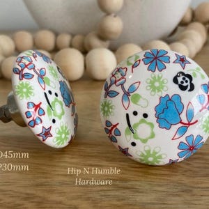 May include: Two ceramic cabinet knobs with a floral design. The knobs are white with blue, red, and green floral patterns. The dimensions are D4.5cm and P3cm. The knobs are on a wooden surface.
