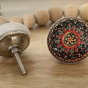 May include: Two ceramic cabinet knobs. One is white with a silver base and a screw. The other is a colourful round knob with a black, red, orange, and green mandala design.