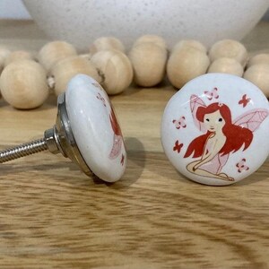 May include: Two white ceramic drawer knobs with a silver metal screw. One knob features a red-haired fairy with pink wings and flowers. The other knob has a floral design.