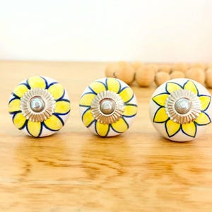 May include: Three ceramic cabinet knobs with a floral design. Each knob is white with yellow petals outlined in blue. A silver-coloured metal centre adds detail. The knobs are displayed on a wooden surface.