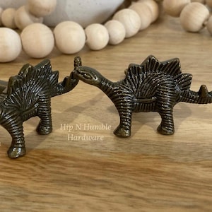 May include: Two bronze dinosaur-shaped cabinet knobs. The knobs are detailed with textured scales and have a screw-on back. The text "Hip N Humble Hardware" is visible in the image.