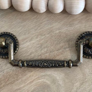 May include: An ornate, brass drawer pull in a vintage style. The pull features a decorative, detailed design with rounded, circular ends and a central bar with a floral pattern.