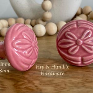 May include: Two pink ceramic cabinet knobs with a floral design. The knobs are 50mm in diameter and 38mm in projection. The text "Hip N Humble Hardware" is visible below the knobs.