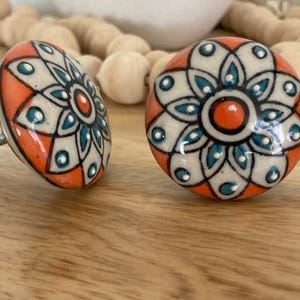 May include: Two ceramic drawer knobs with a white background and a floral design in shades of orange, teal, and black.