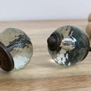 May include: Two faceted glass cabinet knobs with bronze-coloured hardware. The knobs have a clear, textured appearance and are mounted on threaded rods. The hardware includes a washer and nut. The knobs are on a wooden surface.