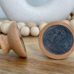 May include: Two wooden cabinet knobs with a light wood finish and a dark grey centre. One knob is lying on its side, showing the screw. The other knob is upright, displaying the grey centre. Decorative home decor.
