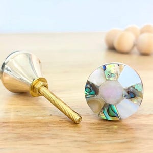 May include: Two decorative cabinet knobs. One has a gold-coloured base and a silver-coloured top. The other is round with a mosaic design of white, iridescent, and pastel-coloured pieces. The knobs are on a wooden surface.