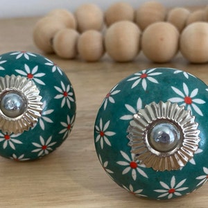 May include: Two teal blue ceramic cabinet knobs with white and red daisy patterns and silver metal accents.