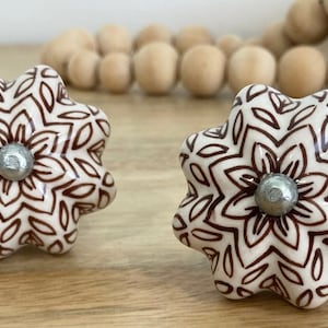 May include: Two white ceramic cabinet knobs with a brown floral design. The knobs have a silver metal accent in the centre.