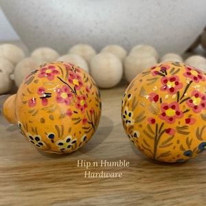 May include: Two round, orange cabinet knobs with a floral design. The knobs are painted with pink, yellow, and blue flowers. The text "Hip n Humble Hardware" is visible below the knobs.