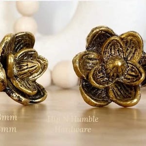 May include: Two gold-toned metal flower-shaped cabinet knobs. The knobs have a detailed, textured design. The text "Hip N Humble Hardware" is visible below the knobs.