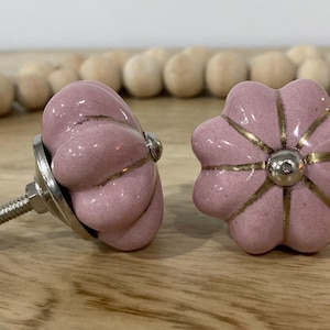 May include: Two pink ceramic cabinet knobs with gold accents. The knobs are shaped like flowers with five petals. The knobs have a silver metal screw on the back.