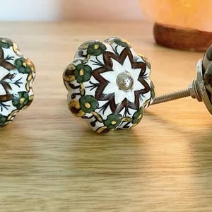 May include: Three ceramic cabinet knobs with a white background and intricate brown, green, and gold floral patterns. The knobs are round and have a star-shaped design in the centre.